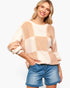 A woman wearing a chunky knit sweater with a large checkered pattern in beige and cream, paired with blue denim shorts, against a plain white background.