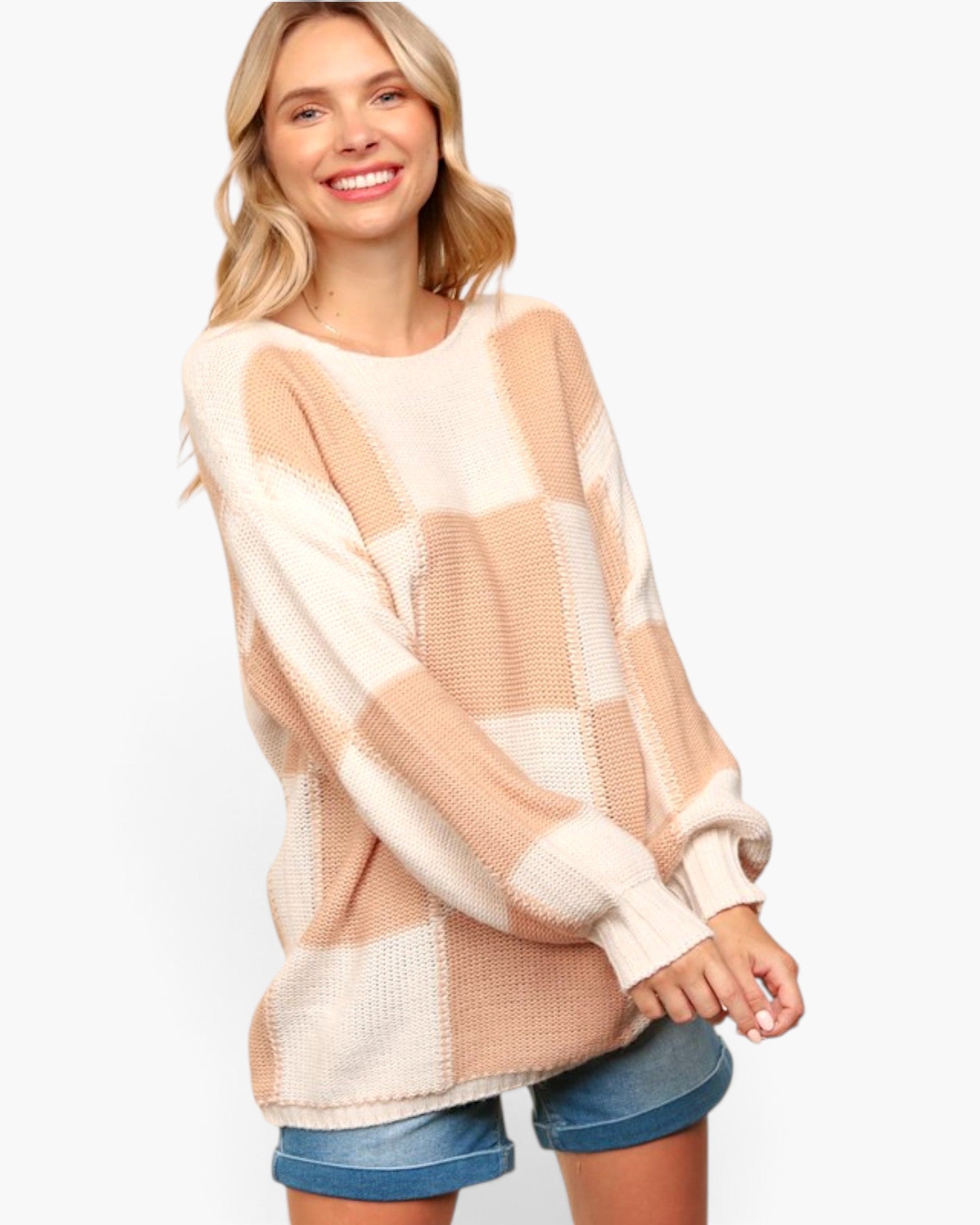 A woman wearing a chunky knit oversized sweater with a large checkered pattern in cream and light tan, paired with denim shorts, against a plain white background.