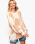 A woman wearing a chunky knit oversized sweater with a large checkered pattern in cream and light tan, paired with denim shorts, against a plain white background.