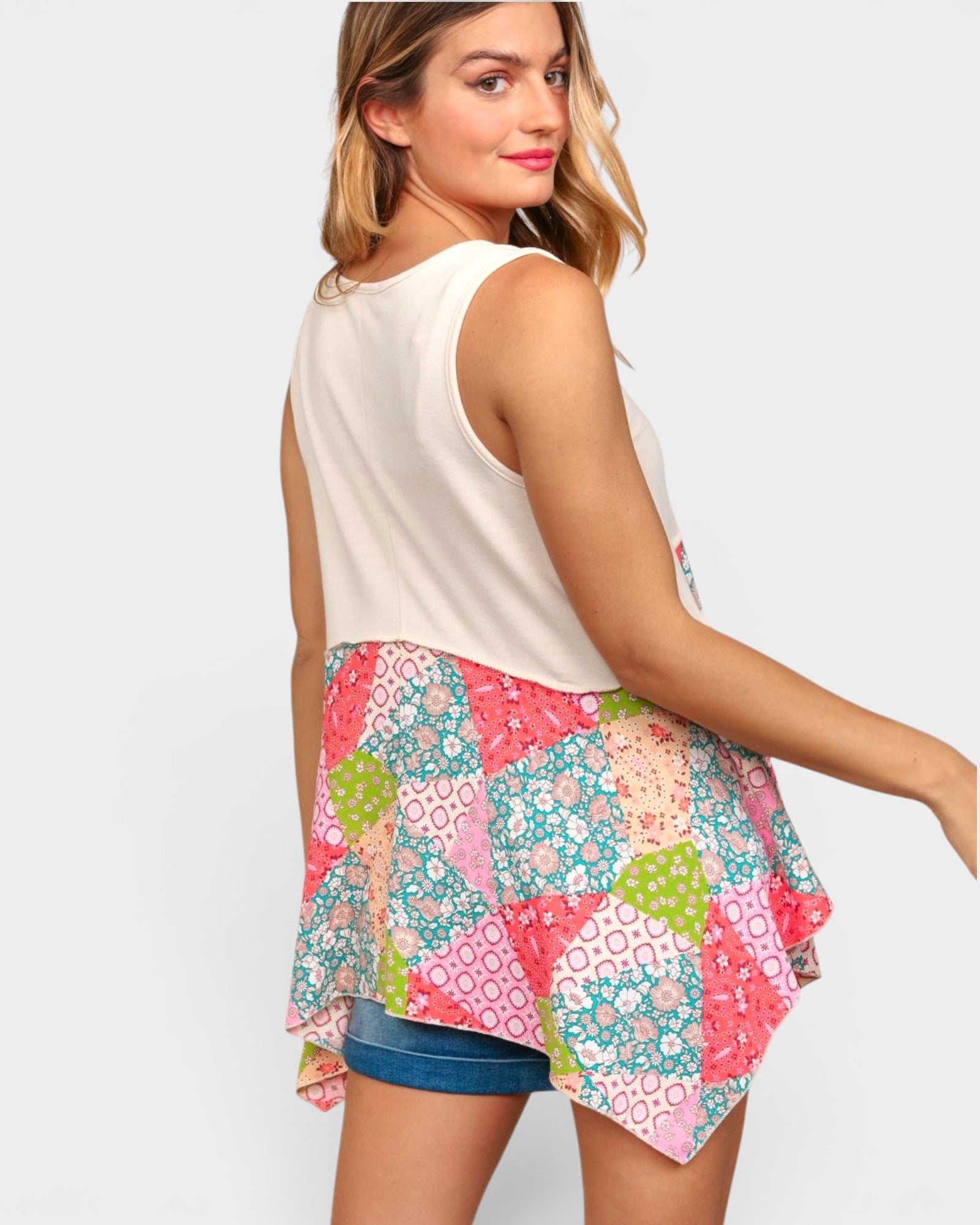 A woman wearing a sleeveless asymmetrical top with a cream upper section and a colorful patchwork floral skirt, paired with denim shorts, shown from a three-quarters back view against a light gray background.