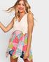 A woman wearing a sleeveless high-low top with a solid cream upper section and a colorful patchwork floral skirt in pink, green, and blue, paired with denim shorts against a light gray background.