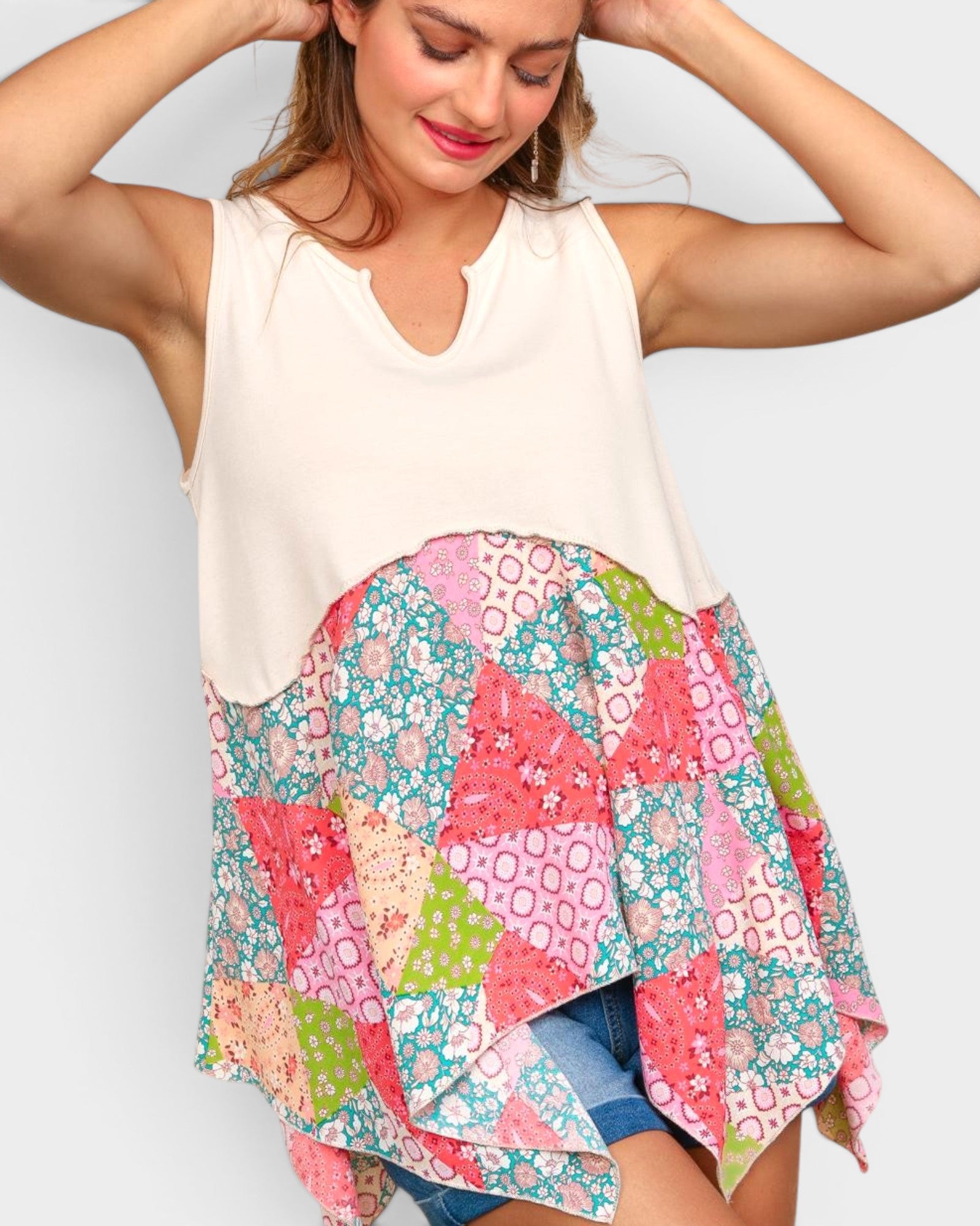 A woman wearing a sleeveless high-low top with a solid cream upper section and a colorful patchwork skirt featuring pink, green, and blue floral patterns, paired with denim shorts against a light gray background.