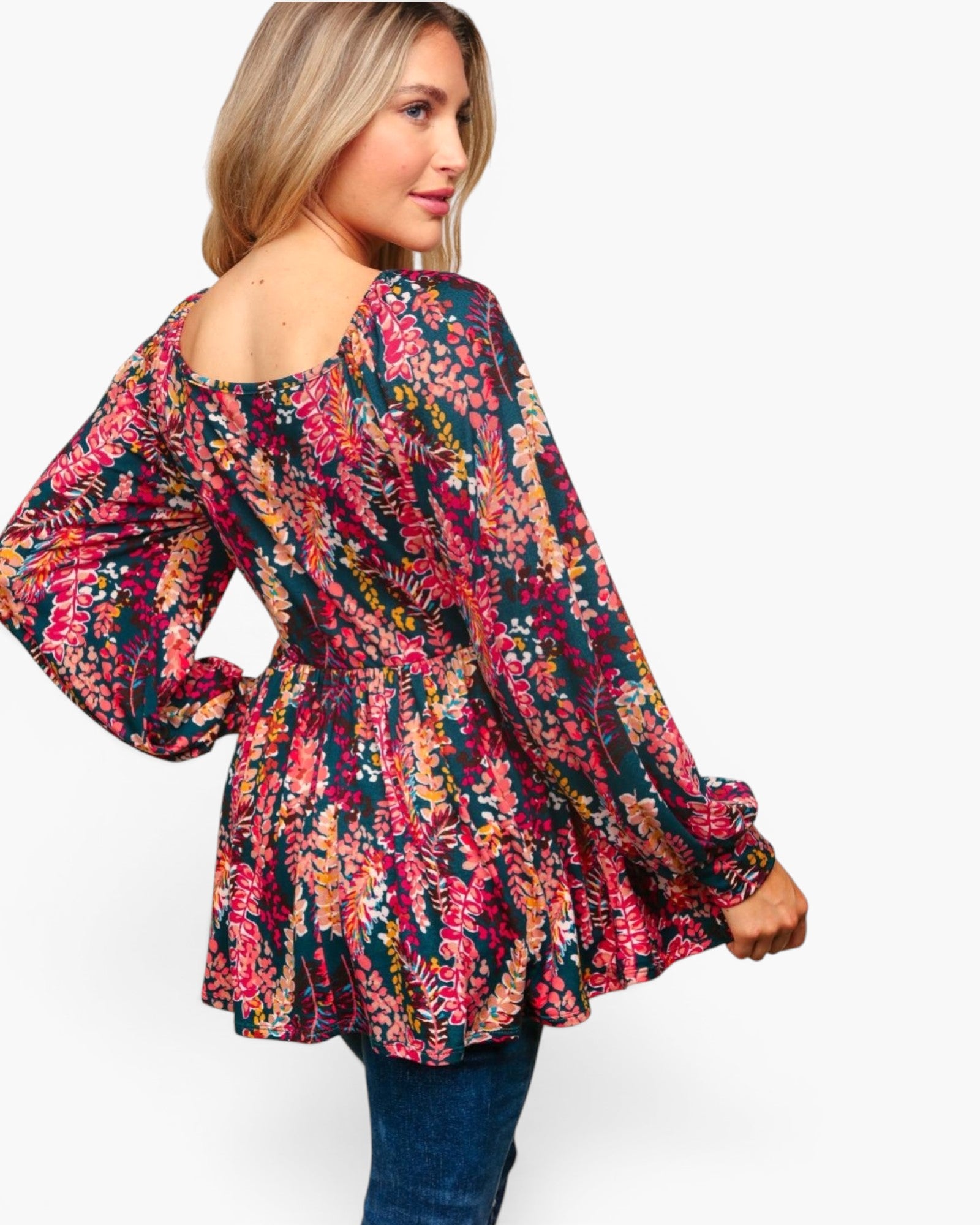 A woman wearing a long-sleeve peplum top with a dense multicolor floral and botanical print, paired with blue jeans, shown from a three-quarters back view against a plain white background.