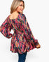 A woman wearing a long-sleeve peplum top with a dense multicolor floral and botanical print, paired with blue jeans, shown from a three-quarters back view against a plain white background.