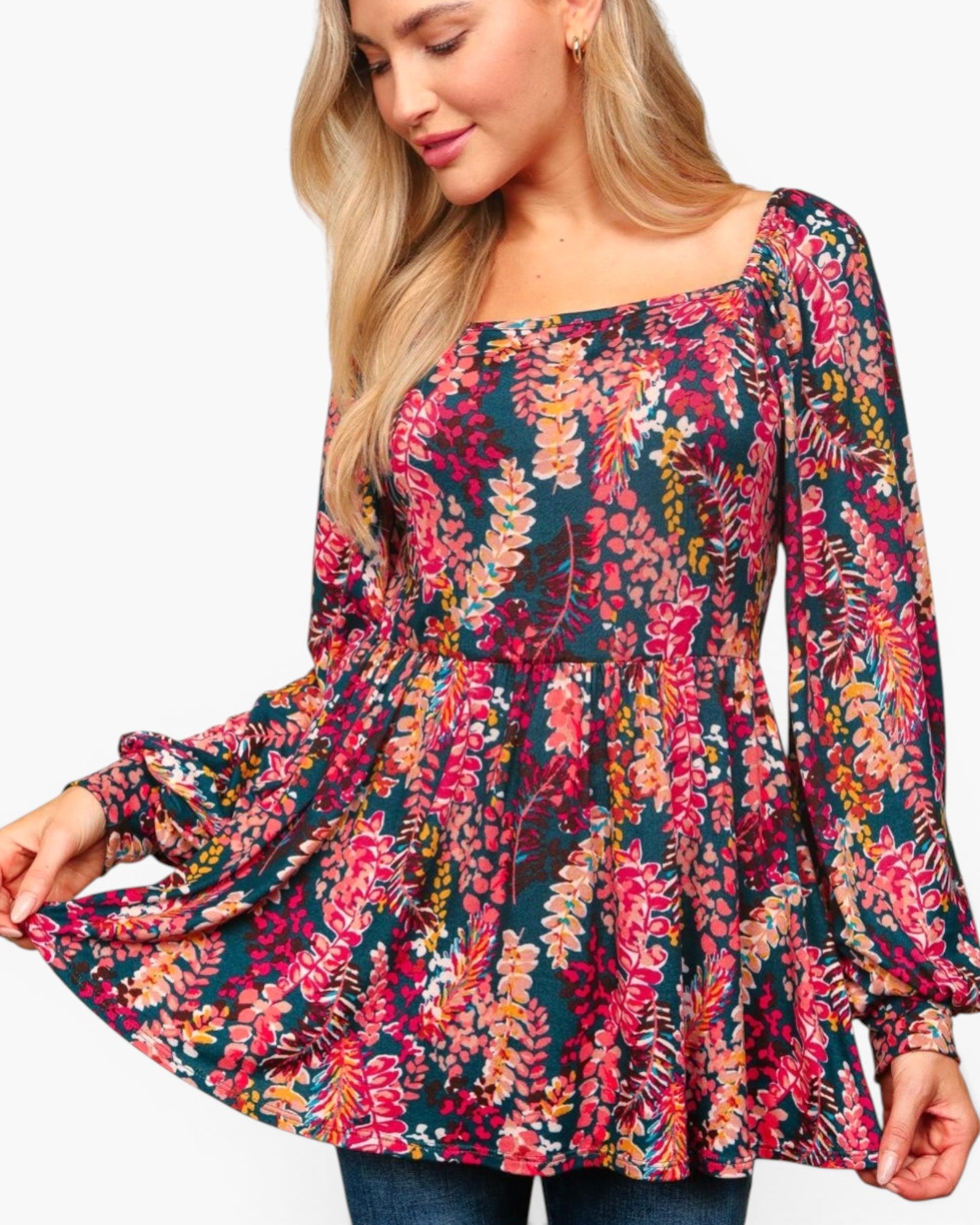 A woman wearing a long-sleeve peplum top with a dense multicolor floral print on a dark teal background, paired with blue jeans against a plain white studio background.