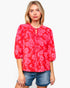 A woman modeling a red and pink floral patterned blouse with a tie-neck detail, paired with denim shorts, against a plain light gray background.