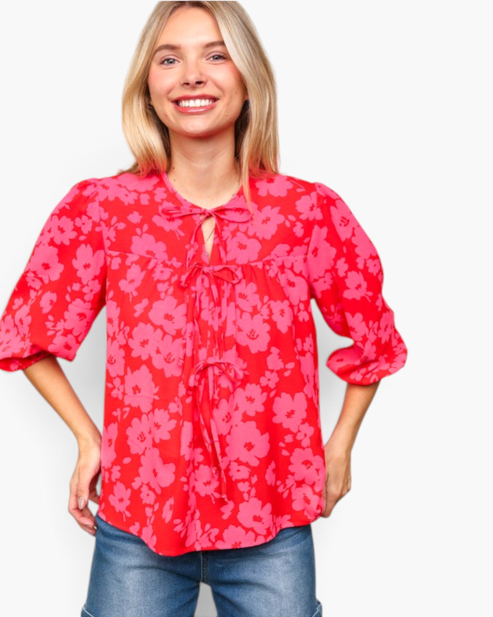 A smiling blonde woman wearing a red and pink floral print blouse with three-quarter sleeves and a tie-neck detail, paired with blue jeans, against a plain light gray background.