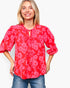 A smiling blonde woman wearing a red and pink floral print blouse with three-quarter sleeves and a tie-neck detail, paired with blue jeans, against a plain light gray background.