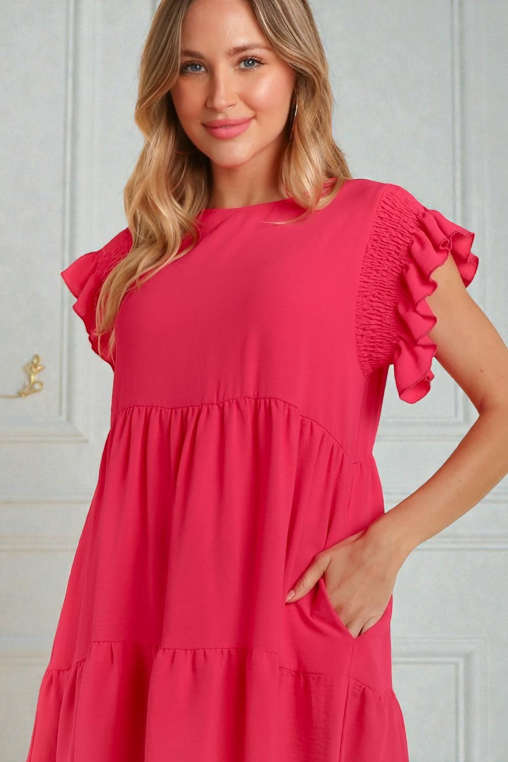 Woman wearing a pink dress with ruffled sleeves against a white wall.