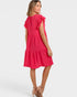A woman modeling a bright pink tiered dress with ruffled cap sleeves, shown from a back-three-quarter angle against a light gray studio background.