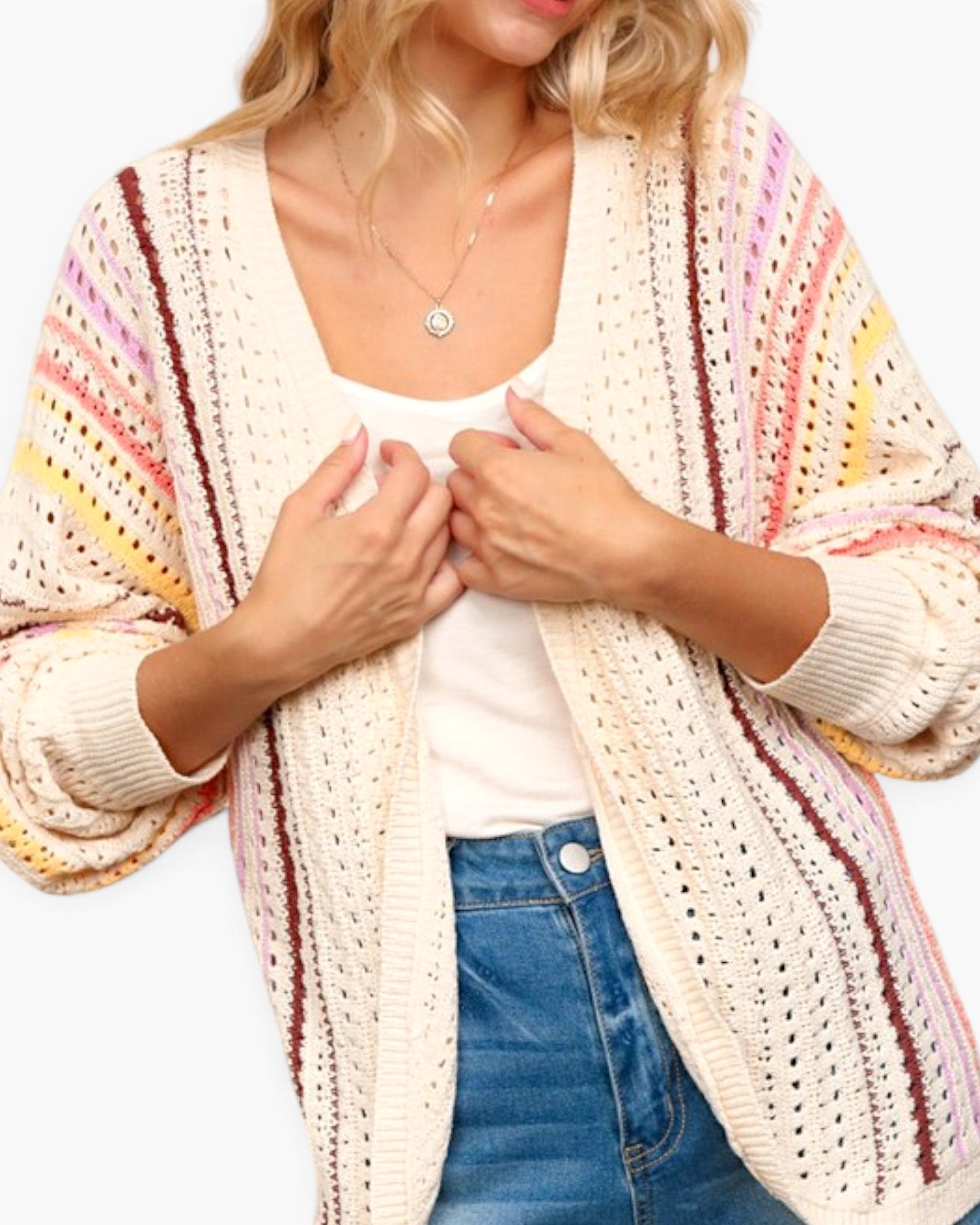 A woman wearing a cream open-front knit cardigan with vertical stripes in pink, yellow, and brown, layered over a white tank top and blue jeans.