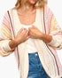 A woman wearing a cream open-front knit cardigan with vertical stripes in pink, yellow, and brown, layered over a white tank top and blue jeans.
