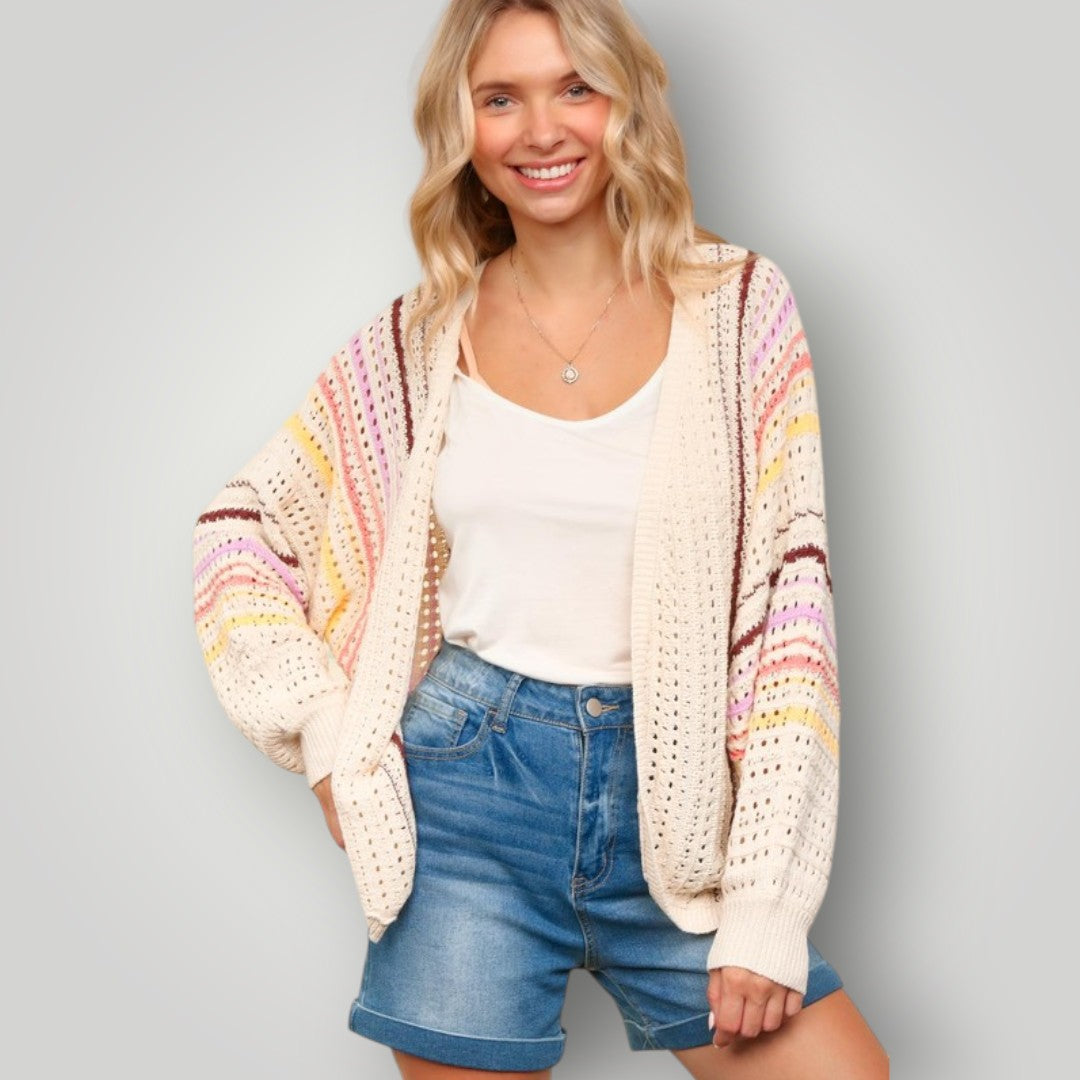 Woman wearing a colorful crochet cardigan over a white top and blue shorts on a gray background