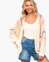 A smiling blonde woman models a cream open-front knit cardigan with colorful pastel stripes in pink, yellow, and blue, worn over a white tank top and blue denim shorts against a plain white background.