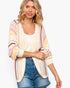 A woman wearing a cream open-front knit cardigan with multicolor striped patterns on the sleeves and sides, layered over a white tank top and blue denim shorts, against a plain light gray background.