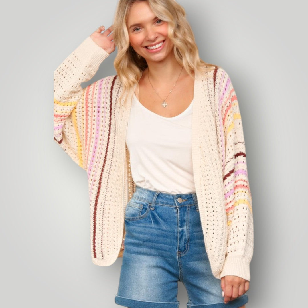 Woman wearing a colorful crochet cardigan over a white shirt and blue jeans on a gray background