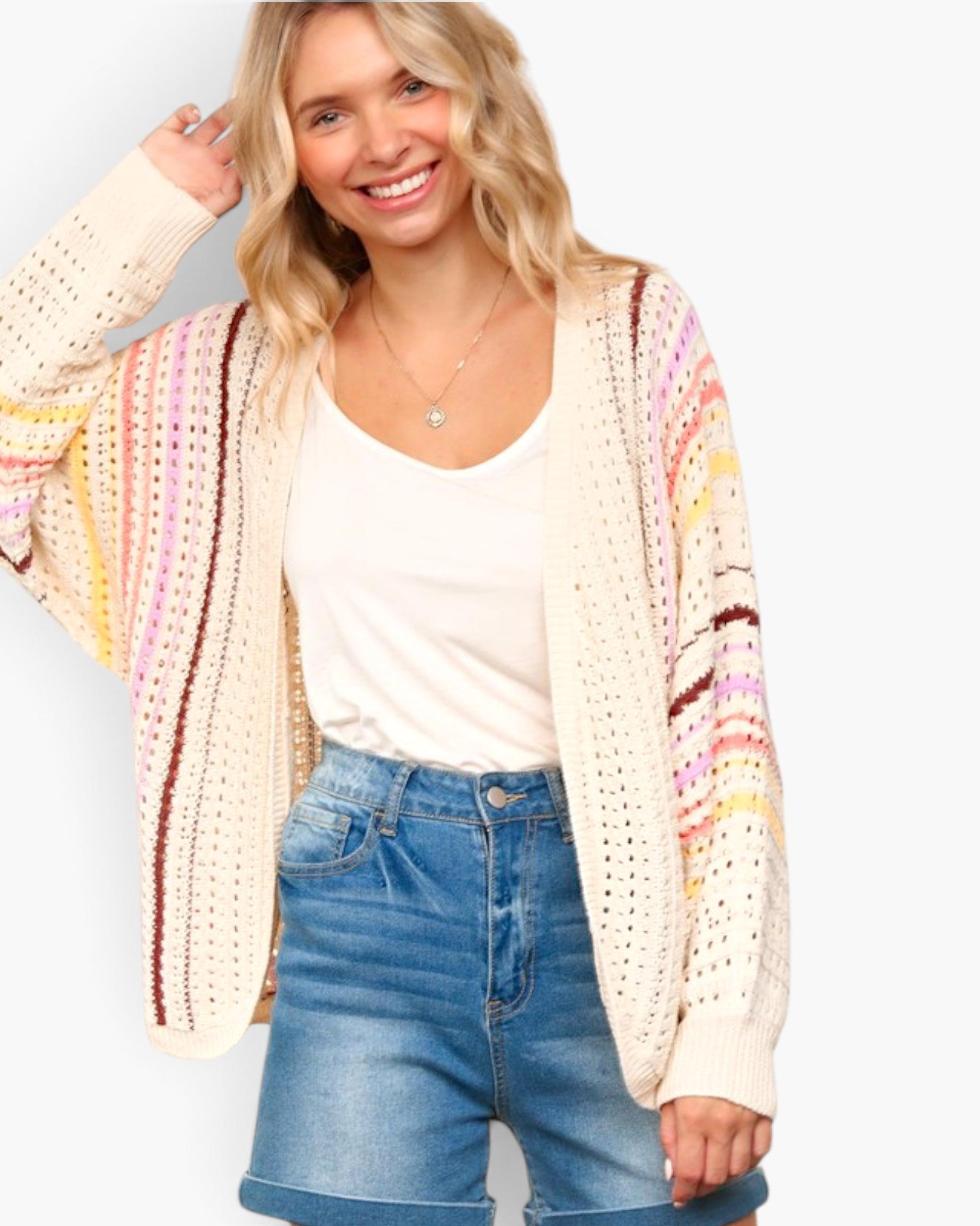 A smiling blonde woman wearing a cream open-front knit cardigan with vertical multicolor stripes in pink, yellow, and brown, layered over a white tank top and blue denim shorts, against a light gray studio background.