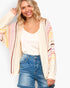 A smiling blonde woman wearing a cream open-front knit cardigan with vertical multicolor stripes in pink, yellow, and brown, layered over a white tank top and blue denim shorts, against a light gray studio background.