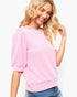 A woman wearing a light pink ribbed knit top with short puff sleeves and a crew neck, paired with blue jeans, against a plain white background.