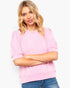 A woman with blonde hair wearing a light pink ribbed short-sleeve top and blue jeans, standing against a plain white background.
