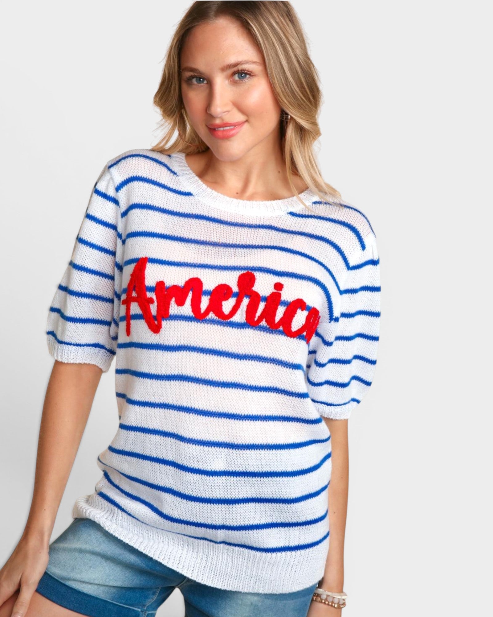 A woman wearing a white short-sleeve knit sweater with blue horizontal stripes and a red 'America' script across the chest, paired with blue denim shorts, against a light gray studio background.