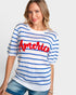 A woman wearing a white short-sleeve knit sweater with blue horizontal stripes and a red 'America' script across the chest, paired with blue denim shorts, against a light gray studio background.