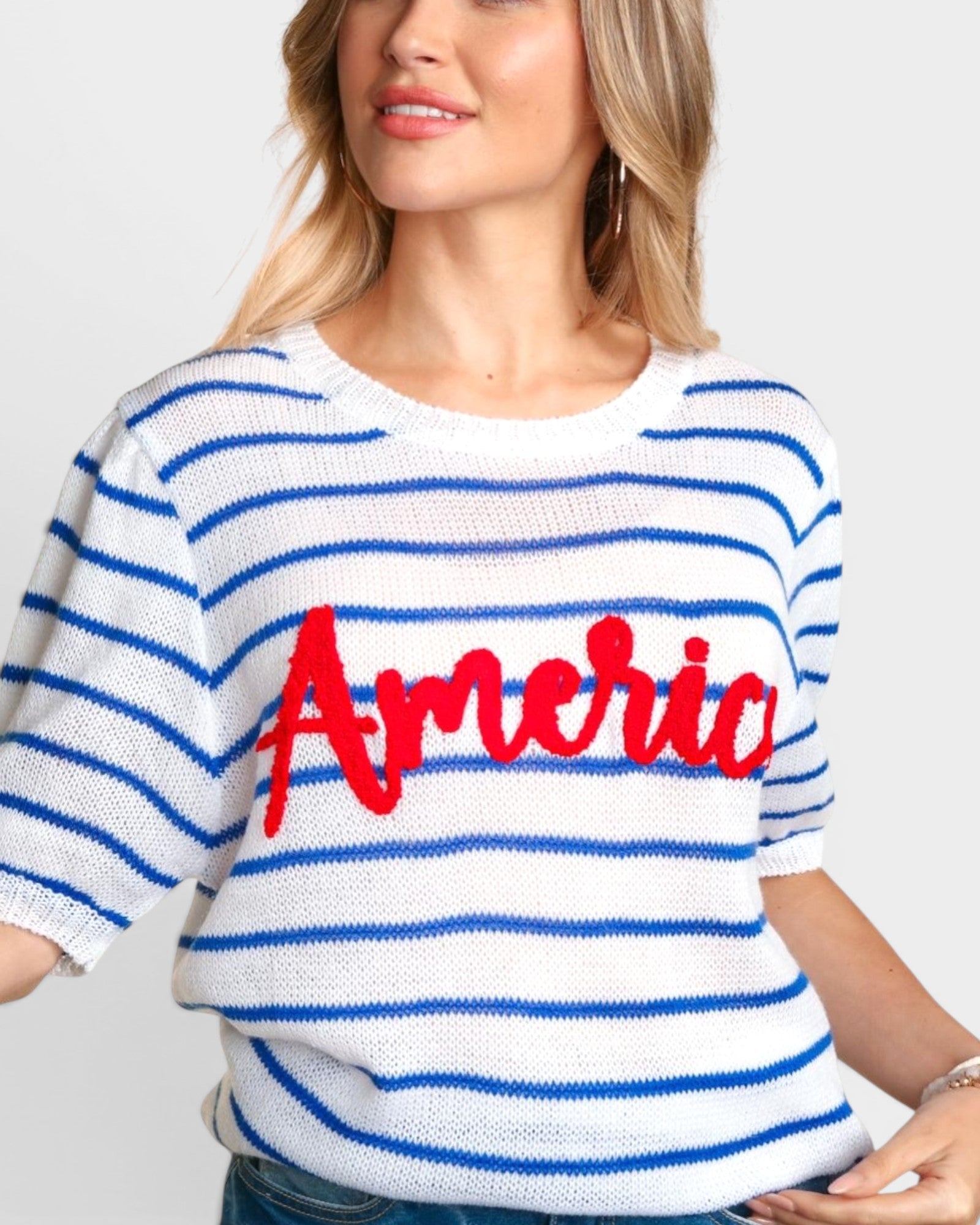 A woman wearing a white knit sweater with blue horizontal stripes and a large red 'America' script across the chest, shown against a light gray studio background.