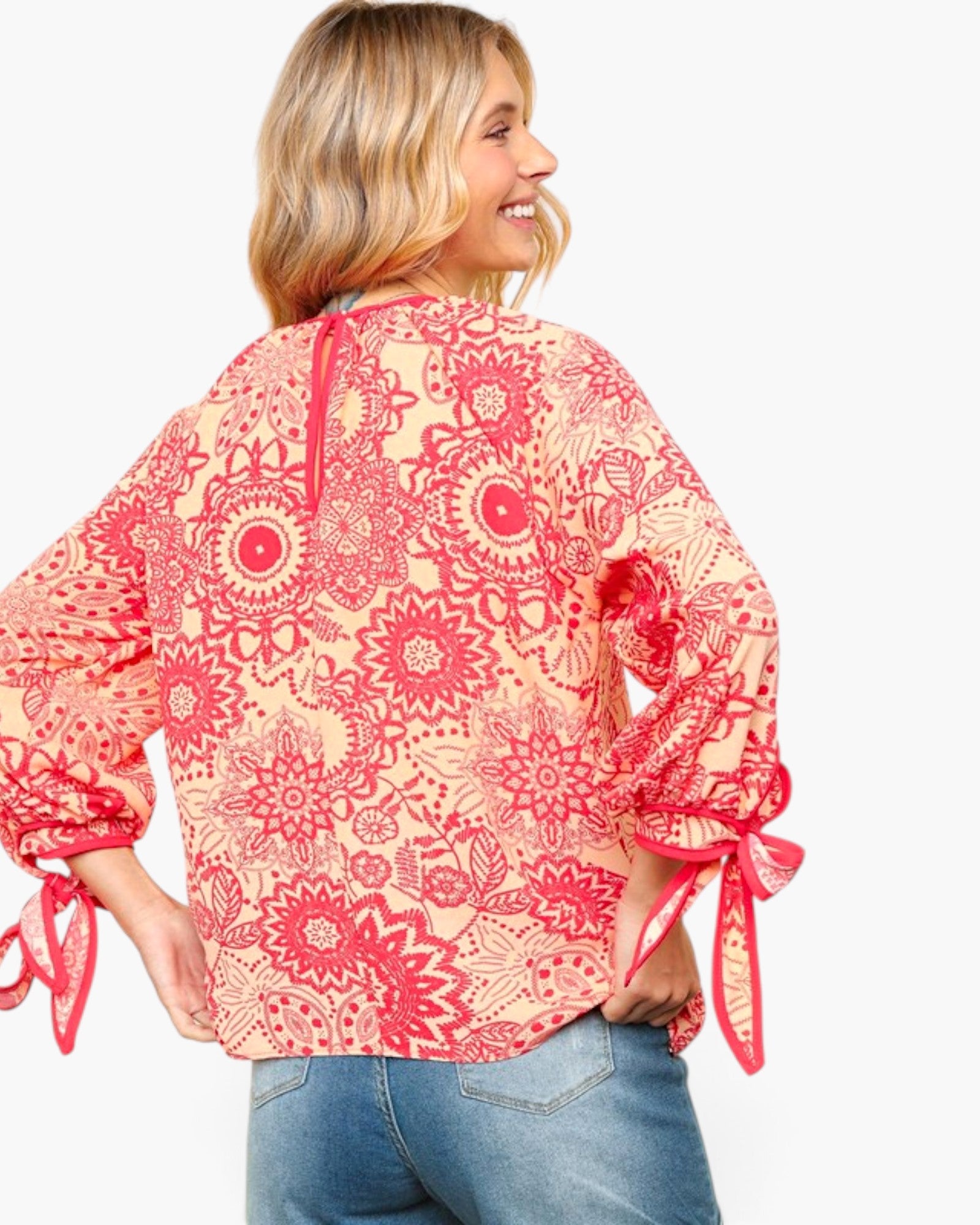 A woman wearing a coral pink and red floral patterned blouse with tie details on the sleeves, shown from the back against a light gray background.