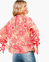 A woman wearing a coral pink and red floral patterned blouse with tie details on the sleeves, shown from the back against a light gray background.