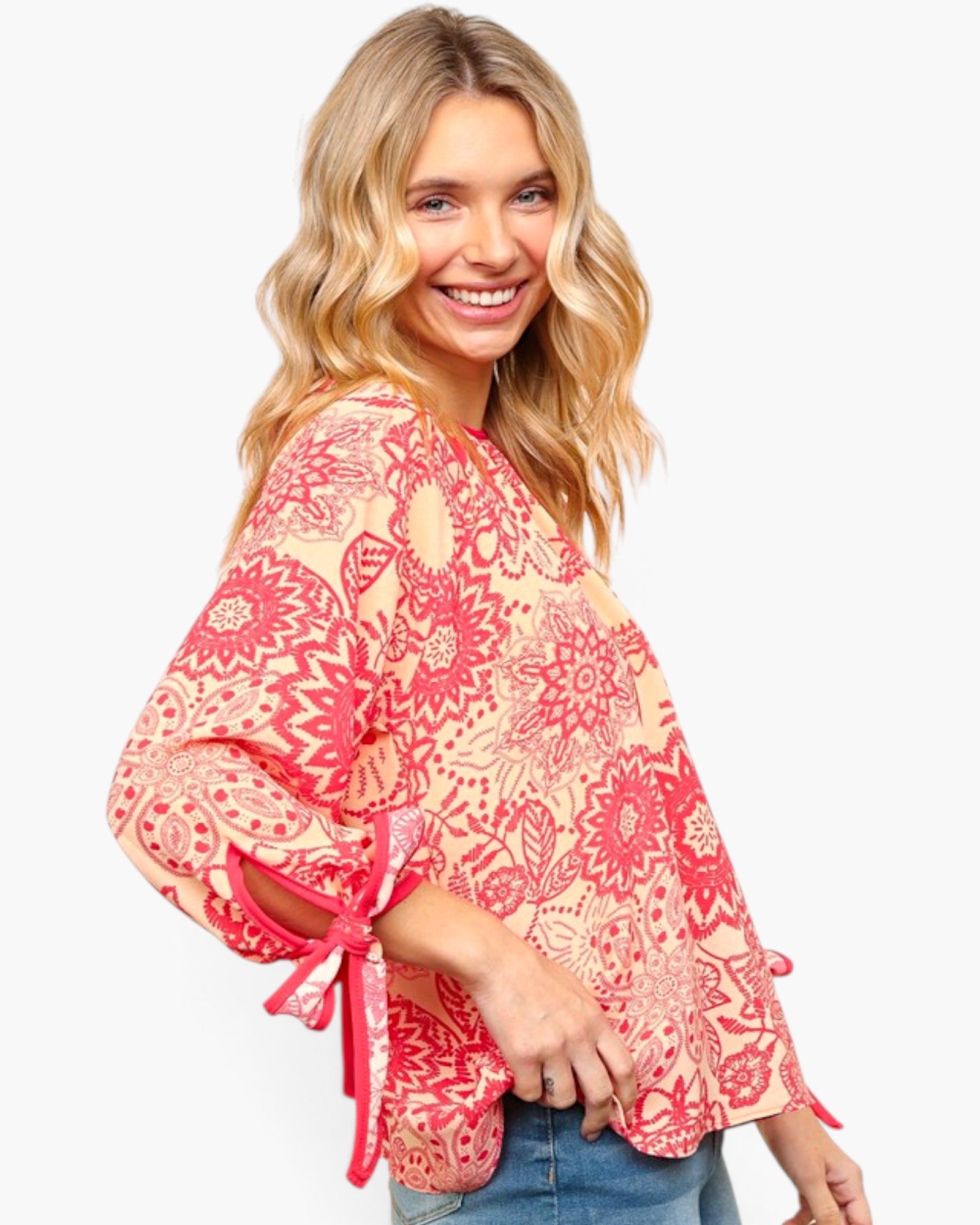 A woman wearing a loose, long-sleeve blouse with a red and beige floral mandala pattern and tie details on the cuffs, paired with blue jeans, against a plain white background.