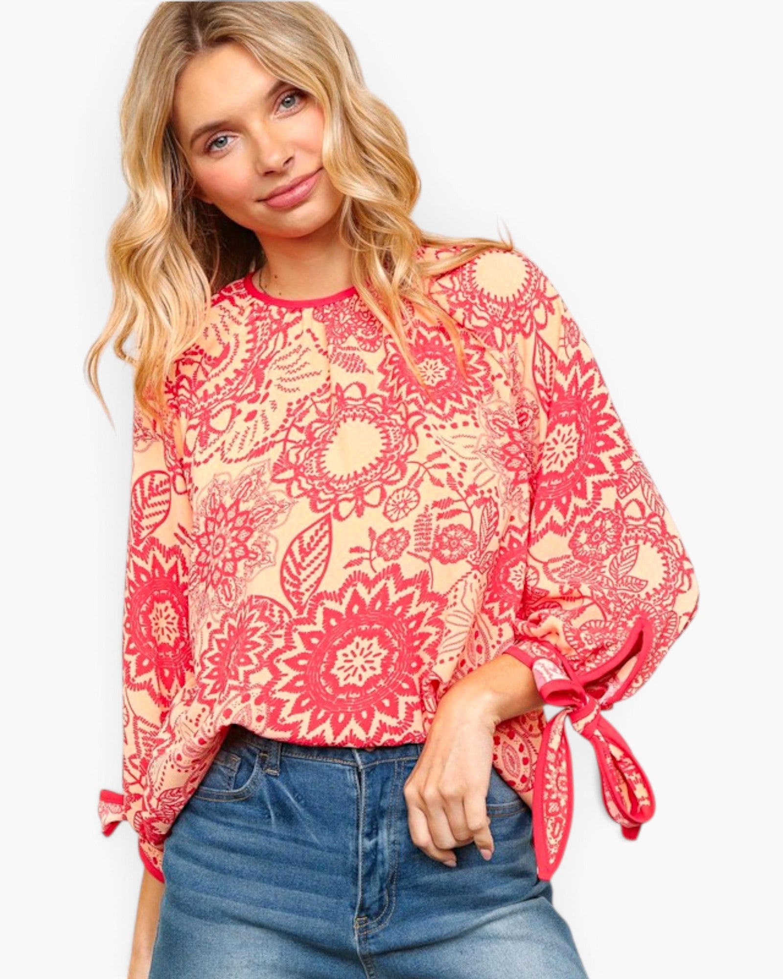 A woman wearing a loose, long-sleeve blouse with a red and beige floral mandala pattern and tie details on the cuffs, paired with blue jeans, against a plain white background.