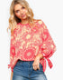 A woman wearing a loose, long-sleeve blouse with a red and beige floral mandala pattern and tie details on the cuffs, paired with blue jeans, against a plain white background.