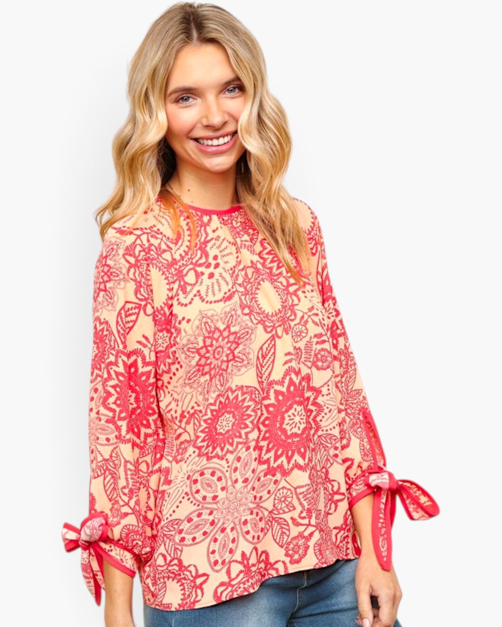 A woman wearing a coral pink and red floral print long-sleeve top with tie details on the cuffs, paired with blue jeans, against a light gray background.