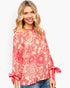 A woman wearing a coral pink and red floral print long-sleeve top with tie details on the cuffs, paired with blue jeans, against a light gray background.