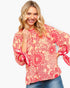 A smiling blonde woman wearing a coral pink and red floral print blouse with tie details on the sleeves, paired with blue jeans, against a plain white background.