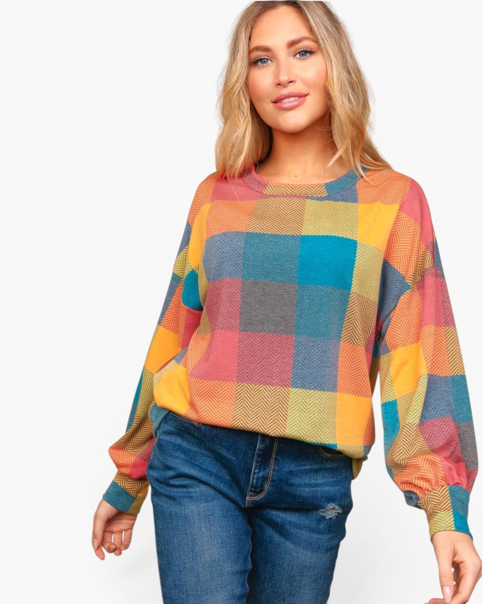 A woman wearing a colorful long-sleeve crewneck sweater with a large plaid pattern in orange, yellow, blue, and pink, paired with blue jeans, against a plain white background.