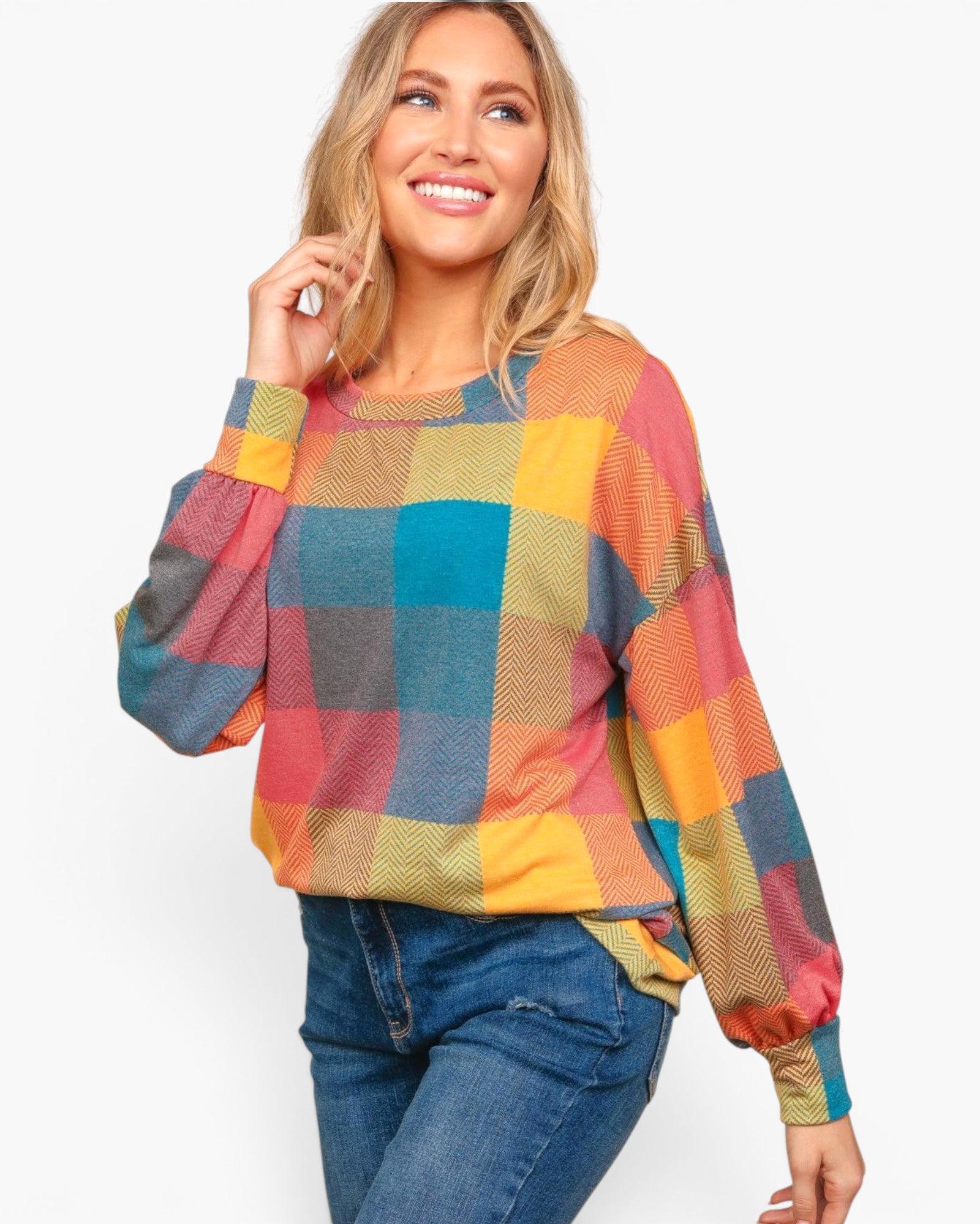 A woman wearing a colorful oversized plaid sweater with blue jeans, photographed against a plain light gray studio background.