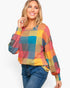 A woman wearing a colorful oversized plaid sweater with blue jeans, photographed against a plain light gray studio background.