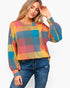 A woman wearing a colorful plaid long-sleeve knit top with blue jeans, posing against a light gray studio background.