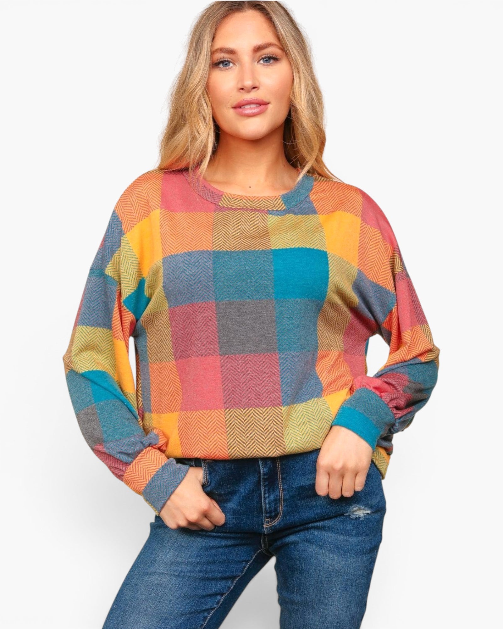A woman wearing a colorful plaid long-sleeve knit sweater and blue jeans, standing against a plain light gray studio background.