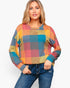 A woman wearing a colorful plaid long-sleeve knit sweater and blue jeans, standing against a plain light gray studio background.