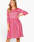 A woman modeling a pink floral fit-and-flare dress with three-quarter sleeves against a plain white studio background.