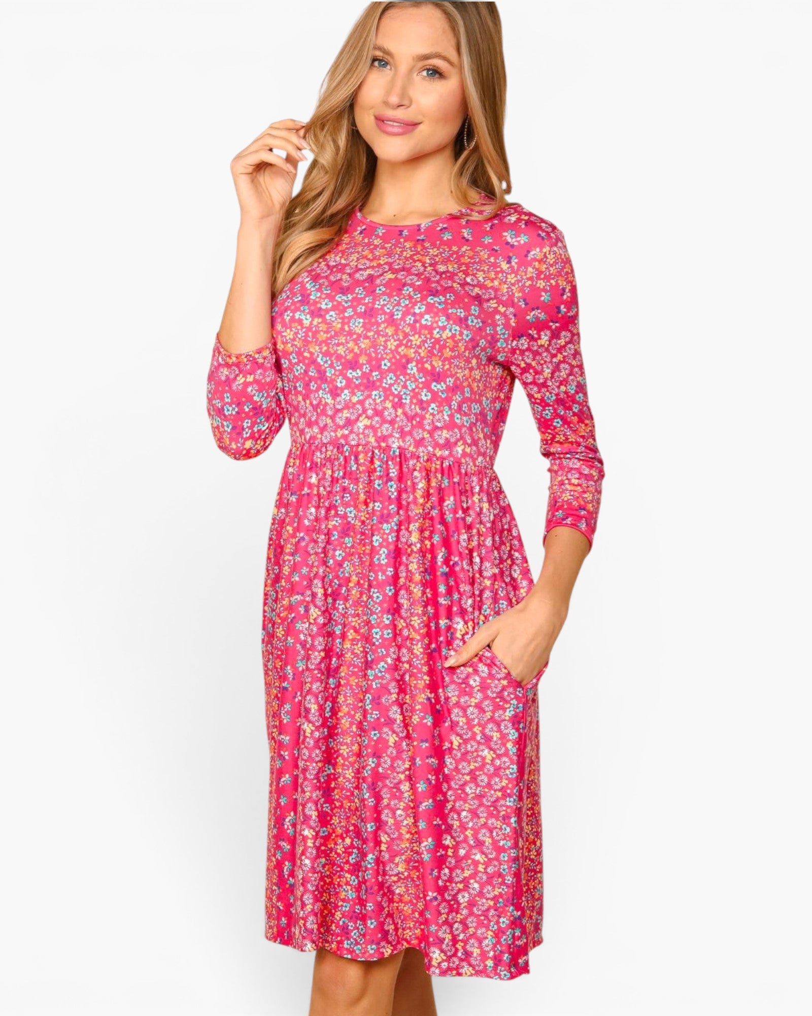 A woman modeling a bright pink three-quarter sleeve midi dress with a dense small floral print in yellow, orange, and white, featuring a gathered waist and side pockets, set against a plain white studio background.
