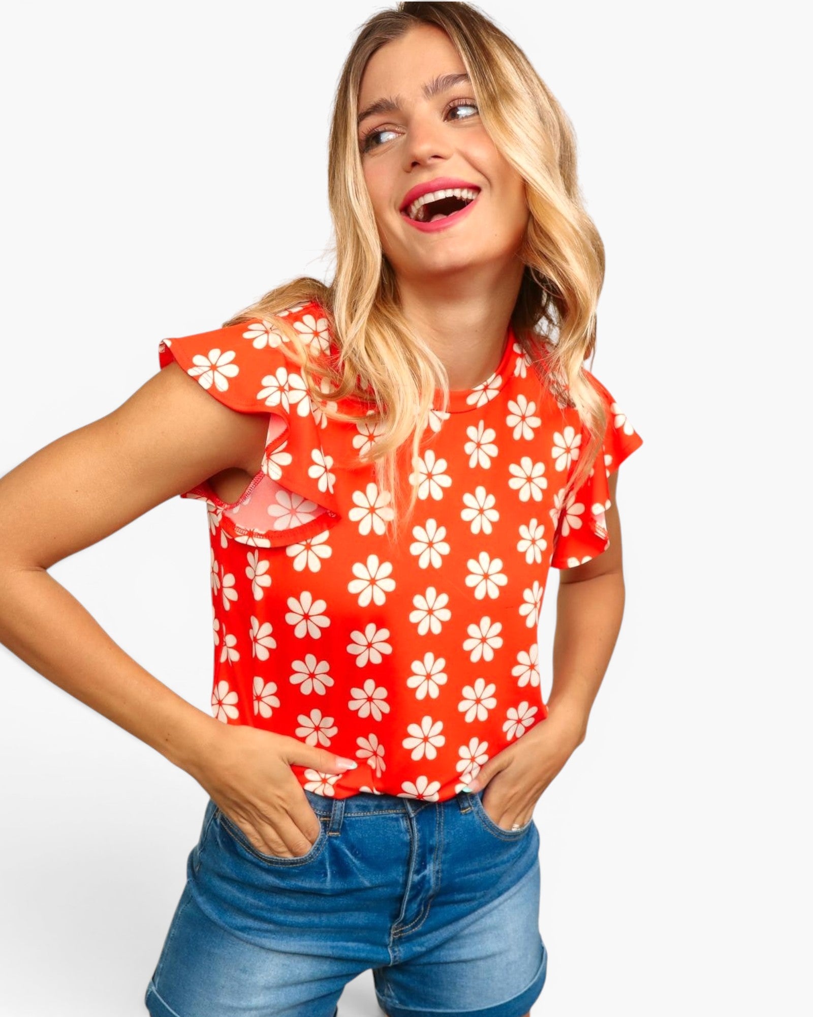 A woman wearing a bright red short-sleeve blouse with a white daisy pattern and ruffled sleeves, paired with blue denim shorts, against a light gray studio background.