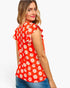 A woman wearing a bright red short-sleeve top with a white daisy floral pattern and ruffled shoulder details, paired with blue jeans, shown from a three-quarters back view against a light gray background.