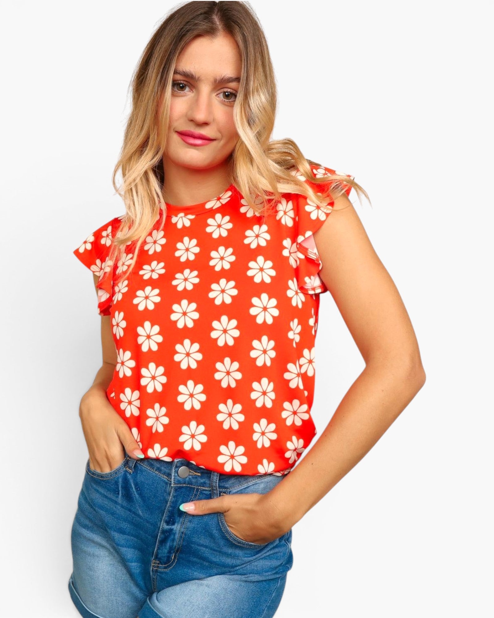 A woman wearing a bright red short-sleeve top with a white daisy floral pattern, paired with blue jeans, against a light gray studio background.