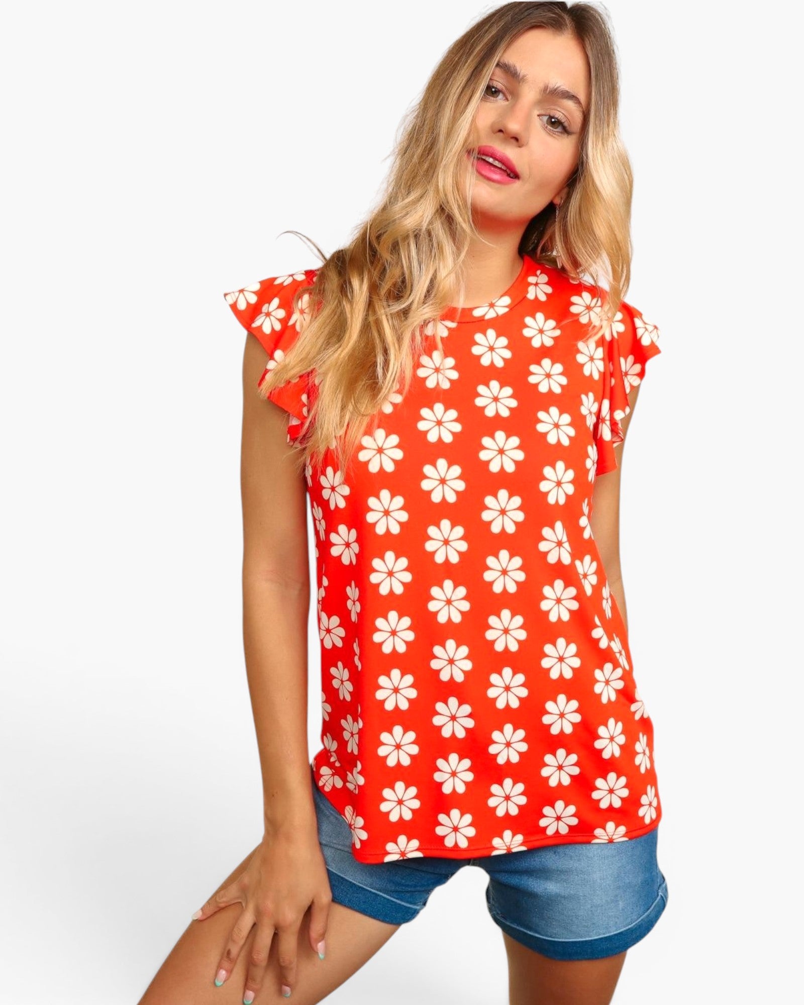 A woman modeling a bright red short-sleeve top with a white daisy floral pattern, paired with denim shorts, against a plain white studio background.