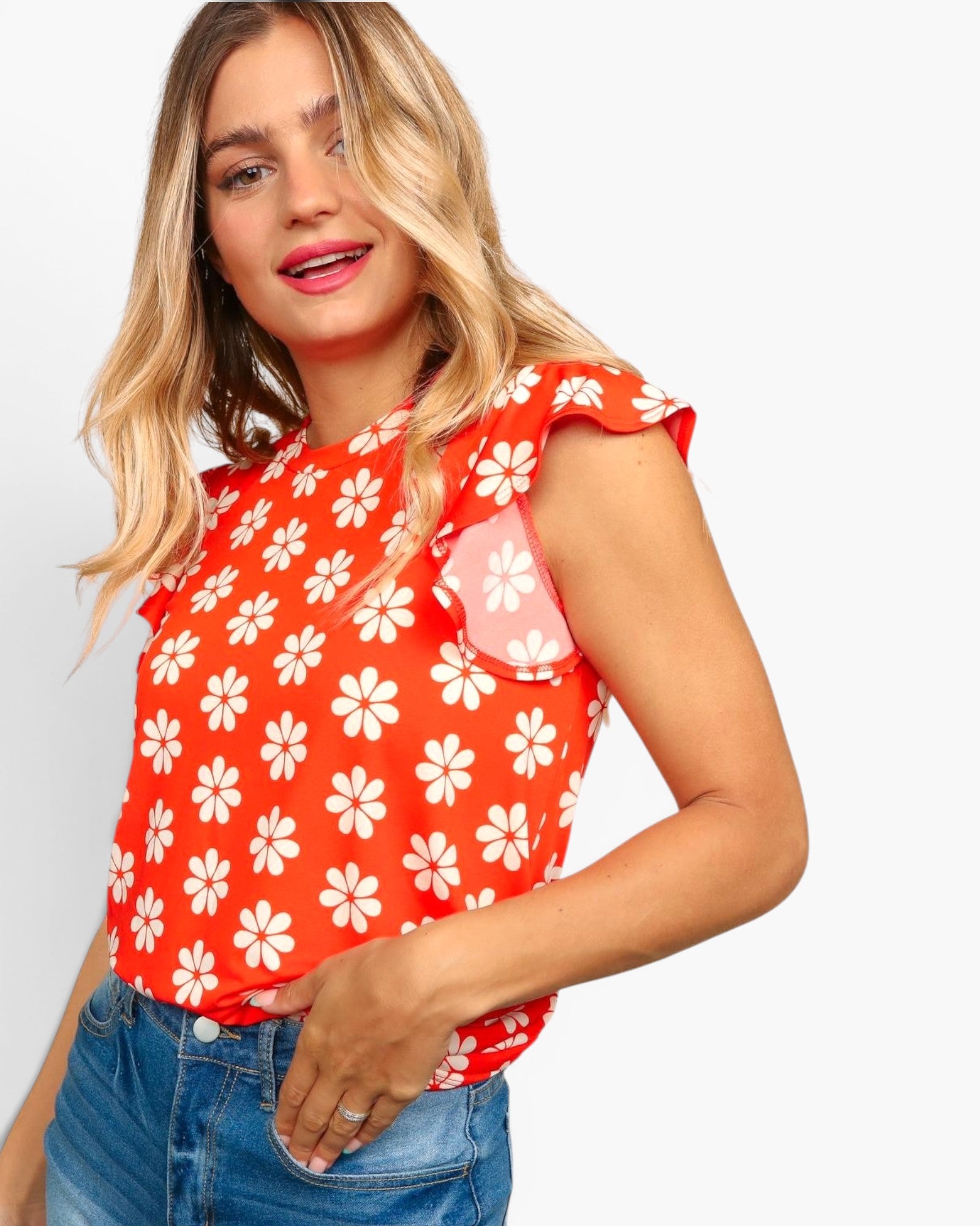 A woman wearing a bright red sleeveless top with a white daisy floral pattern and ruffled cap sleeves, paired with blue jeans, against a light gray background.