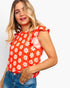 A woman wearing a bright red sleeveless top with a white daisy floral pattern and ruffled cap sleeves, paired with blue jeans, against a light gray background.