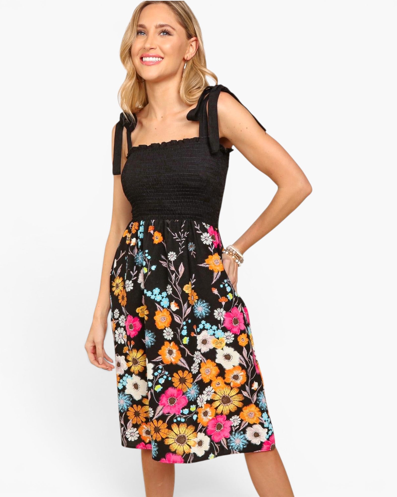 A woman modeling a sleeveless midi dress with a black smocked bodice and a black skirt featuring a vibrant multicolor floral print, set against a plain white background.